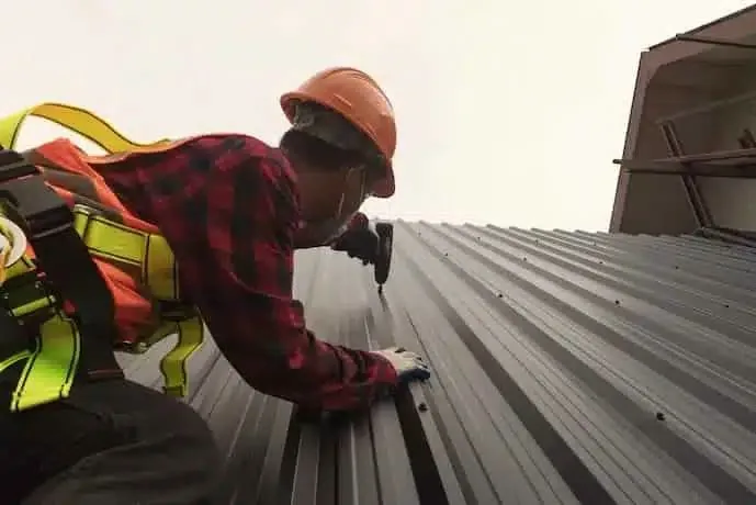 Profissional de capacete e cinto de segurança parafusando telha metálica trapezoidal na cobertura de um galpão
